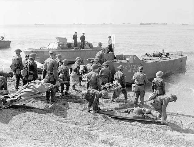 Black and white photograph. On a rocky beach, men in battle dress stand with their backs to the camera looking at the two small boats on the shoreline. Some hold stretchers with men on them. They wait to board the stretchers on the boats.
